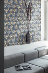 A luxurious, maximalist wallpaper featuring a botanical, Japanese and Arts and Crafts-style pattern in metallics, blue, grey, pink and yellow, with a subtle shimmer, in a modern living room with a grey sofa. Wild Lone's "Berry Galaxy" wallpaper in "Smoky Grey".