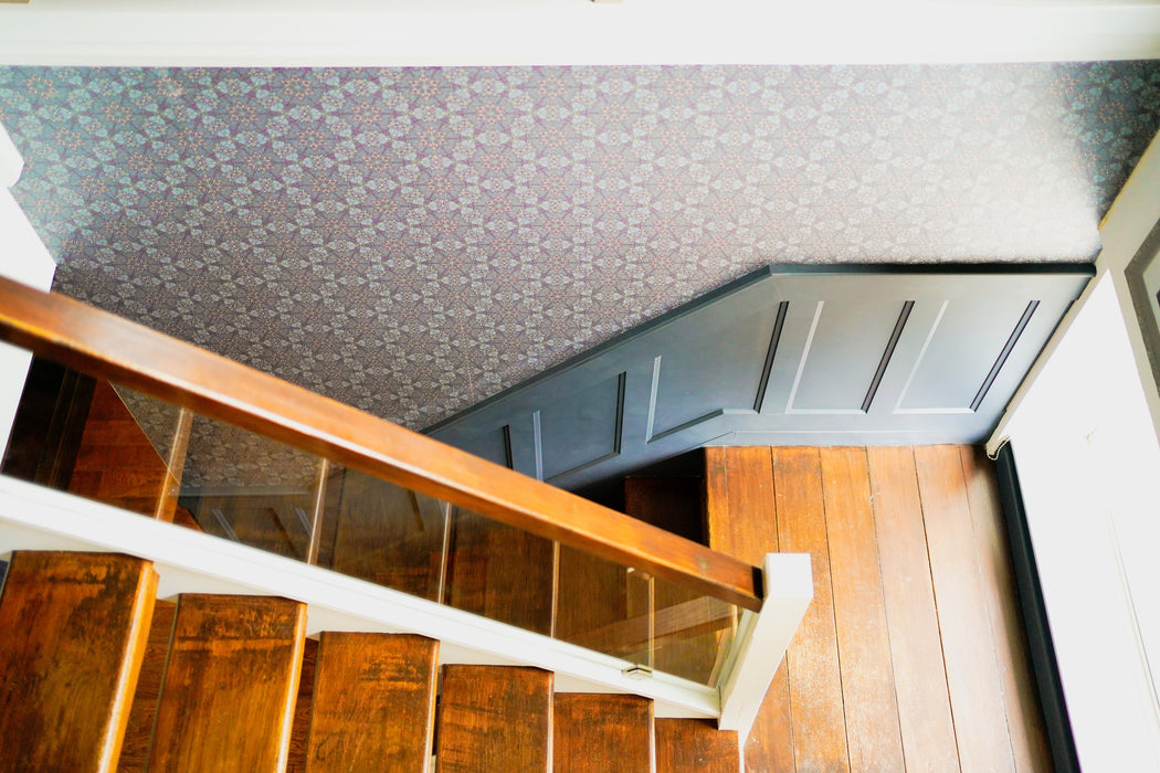 A bold and moody wallpaper in blue, pink and gold, with an intricate, botanical pattern inspired by Art Nouveau and Japanese design, above blue wainscoting in a stairwell in a period home. Wild Lone's "Greenhouse" wallpaper in "Ornamental Blue".