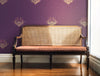 A moody, jewel tone wallpaper featuring fantastical bees, in a period house, in the hallway by a large window with a long, antique, occasional chair with a cane back (Louis XVI style). Wild Lone's "Night Flight" wallpaper in "Purple Jewel", featuring a deep purple background and antique gold and magenta bees.